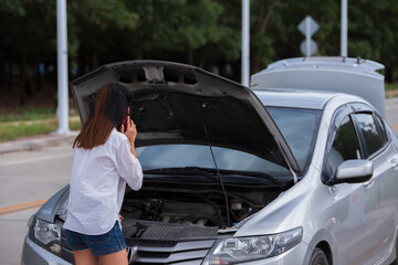Young woman using mobile phone while looking at broken down car on street.