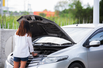 Young woman using mobile phone while looking at broken down car on street.