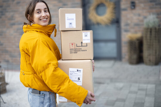 Cheerful woman in bright jacket with parcels in front of her house, coming home with goods purchased online. Shopping online and delivering goods concept