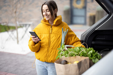 Woman arrives home with a groceries, standing with phone near the car trunk. Shopping healthy food...