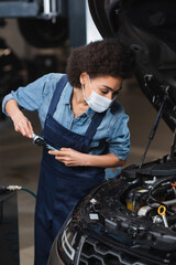 focused young african american mechanic in protective mask holding wrench and working with car motor in garage