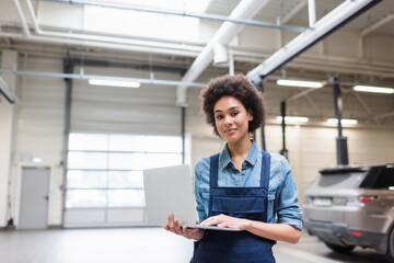 cheerful young african american mechanic standing with laptop and looking at camera in garage