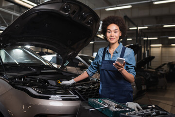 smiling young african american mechanic standing with cellphone near car with open hood in garage