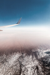 View from the window of an airplane with a wing and a folded terrain covered with snow and glaciers in the Iranian highlands