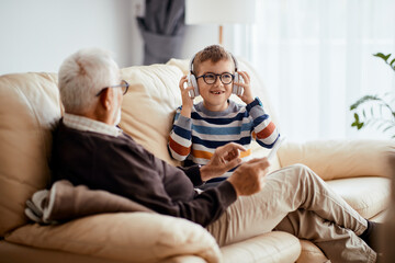 Happy kid listening music over headphones while spending time with grandfather at home.