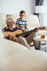 Happy senior man playing acoustic guitar to his grandson at home.