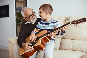 Happy kid having fun while grandfather is teaching him to play acoustic guitar at home.