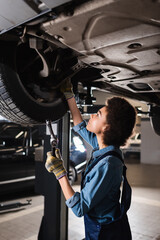 young african american mechanic in overalls holding wrench and repairing wheel of lifted car in garage