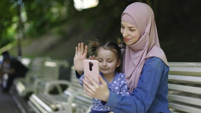 Pretty young Arabian woman in hijab, sitting in the park on the bench together with her adorable little daughter, having fun, waving to phone camera during video call. People, family, technologies