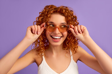 Cute red-haired girl with curls and sunglasses is having fun isolated on purple background.