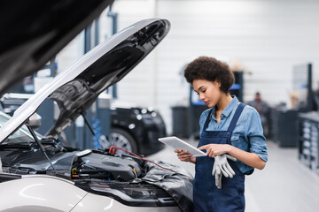 young african american mechanic holding digital tablet near car with open hood in auto repair...