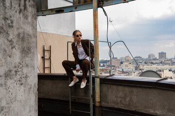 full length of young model in stylish sunglasses and trendy suit sitting near metallic stairs on rooftop