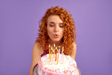 Cute red-haired woman with curls in a shiny dress holds a cake and blows out the candles isolated...