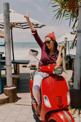 Excited stylish young woman dressed cap and shirt sitting on red bike posing on background of ocean...