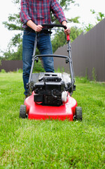 Fototapeta premium Lawn grass mowing. A man in a plaid shirt and blue jeans mows the grass with a lawn mower.