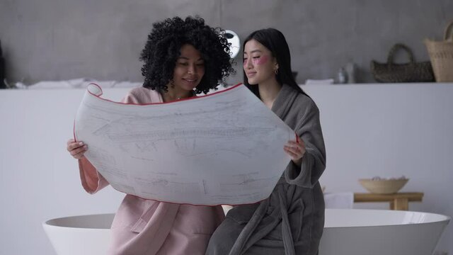 Two Creative Positive Young Women Discussing Architectural Blueprint Laughing Sitting On Bathtub In Bathroom. Charming African American And Asian Roommates Talking Analyzing Project In Home Office