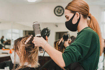 Fototapeta premium Hairdresser in a mask with at work, drying hair in a barbershop, client and stylist in masks.