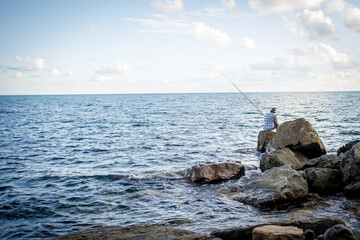 Fototapeta na wymiar A fisherman is fishing in the sea sitting on large boulders.