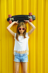 Little cute girl in sunglasses holding skate on yellow background. Summer vibes concept.