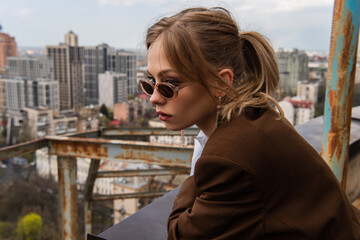 thoughtful woman in stylish sunglasses posing on rooftop with blurred cityscape on background