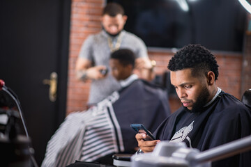 young girl checking cell phone.wait while they start cutting hair in latin hair salon.hairdresser in background cutting boy's hair. .