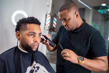 hairdresser cutting the hair of a young man in a latin barber shop.with haircutting machine carefully in front of the mirror.with watch and ring.and led light ring behind the client..