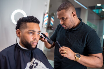 hairdresser cutting the hair of a young man in a latin barber shop.with haircutting machine carefully in front of the mirror.with watch and ring.and led light ring behind the client..