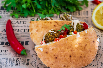 Authentic fresh falafel balls inside of two halves of pita bread sandwich with chopped salad, red hot peppers, lemon and a drizzle of tahini sauce. chickpea falafels in fluffy pita on decorative paper