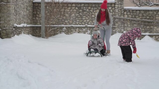 vacation, game, family concept - slo-mo authentic three happy preschool toddler kids siblings with young wonan mom sledding and play with snow. snowfall in cold season weather in winter yard outdoors