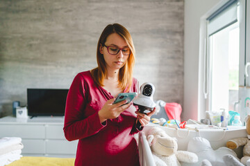 Caucasian woman mother adjusting and setting up Surveillance security camera on baby bed at home in...