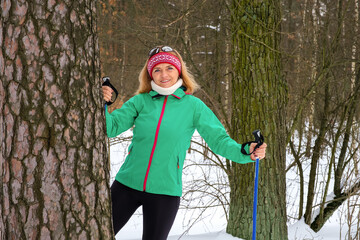 Elderly woman standing with nordic walking poles behind the tree with outstretched arms in winter forest, resting after exercise and looking at camera outdoors with smile.