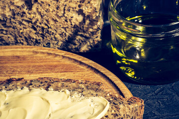Slice of rustic natural yeast-free bread with flax, poppy seeds, sesame seeds, millet, pumpkin and sunflower seeds, spread with butter, on a wooden board, with olive oil in a glass jar