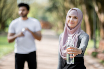 Blur background of young woman in hijab and muslim man standing at green city park with fresh water. Focus on female hand with bottle of water. Refreshing concept.