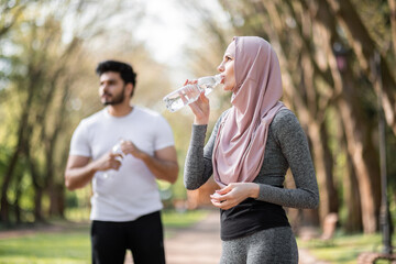 Side view of attractive woman in hijab drinking water while handsome arabian guy standing behind. Young couple in sportswear relaxing after workout on fresh air.