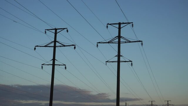 View of a high voltage power line at dawn. Landscape in the early morning overlooking silhouettes of the electricity power supply poles in a plain on clear blue sky background with bit clouds.