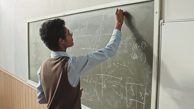 Rear-view medium slowmo of young Mixed-race high school or university student in smart casualwear solving Geometry problem at blackboard