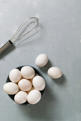 White chicken eggs lie in a gray bowl and whisk on a gray background.