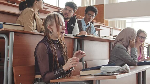 Medium slowmo of young female student with dreadlocks wearing gothic clothes and makeup chewing gum and scrolling through phone getting bored during lesson