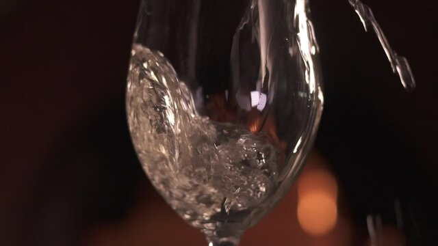 Close-up of a tall transparent glass in which white wine is poured in dark room against the background of the fireplace. The wine spills and drips past the glass. Bokeh fire in the background. 120 fps