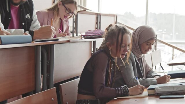 Tilt-down medium slowmo of diverse male and female students sitting in rows by long wooden desks in lecture hall and chatting during lesson