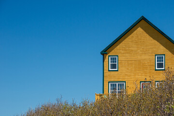 A tall multiple story orange colored wooden building with clapboard siding double hung windows with green and white trim and a steep peaked shingled roof. The background is a sunny blue sky.