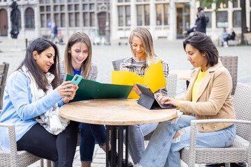 Lifestyle portrait of a diverse multiethnic group of four young smiling businesswomen with smartphones, notes and notebook outside in a cafe at work while having a drink. High quality photo