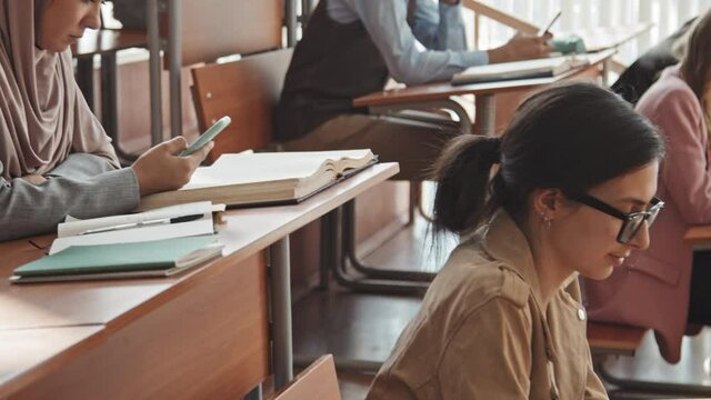 Tilt-up medium slowmo of young multi-ethnic university or high school students sitting in rows by long wooden desks in auditorium and scrolling through smartphones