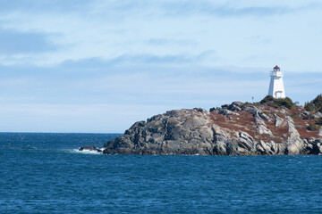 A rugged coastline and cliff in the foreground near a deep blue ocean. It's a bright sunny day with a blue sky and few clouds. There's an island in the background with greenery, buildings, and rock. 