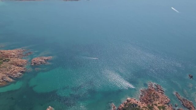 Aerial view of a reddish coastline with some boats crossing a transparent blue sea near Isola Rossa, Sardinia, Italy.