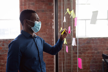 African american businessman wearing face mask brainstorming, reading memo notes on glass wall