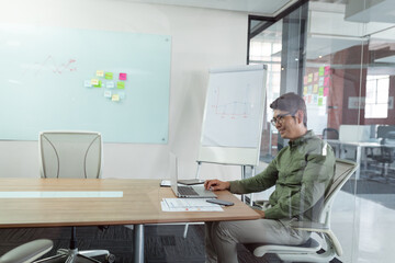 Smiling asian businessman sitting in meeting room using laptop seen through glass wall