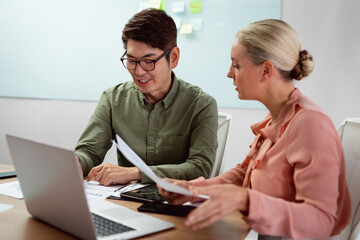 Diverse male and female colleague sitting at table with laptop and paperwork discussing