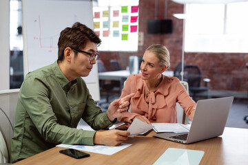 Diverse male and female colleague sitting at table with laptop and paperwork discussing