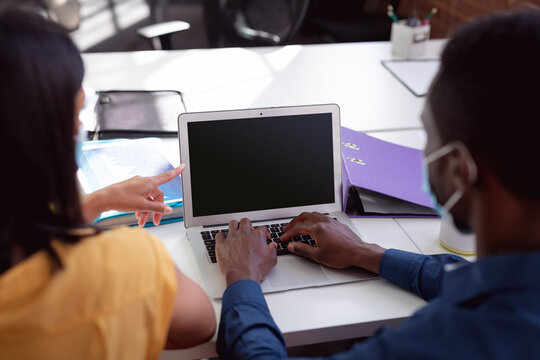 Diverse male and female colleague in face masks sitting at desk discussing and using laptop